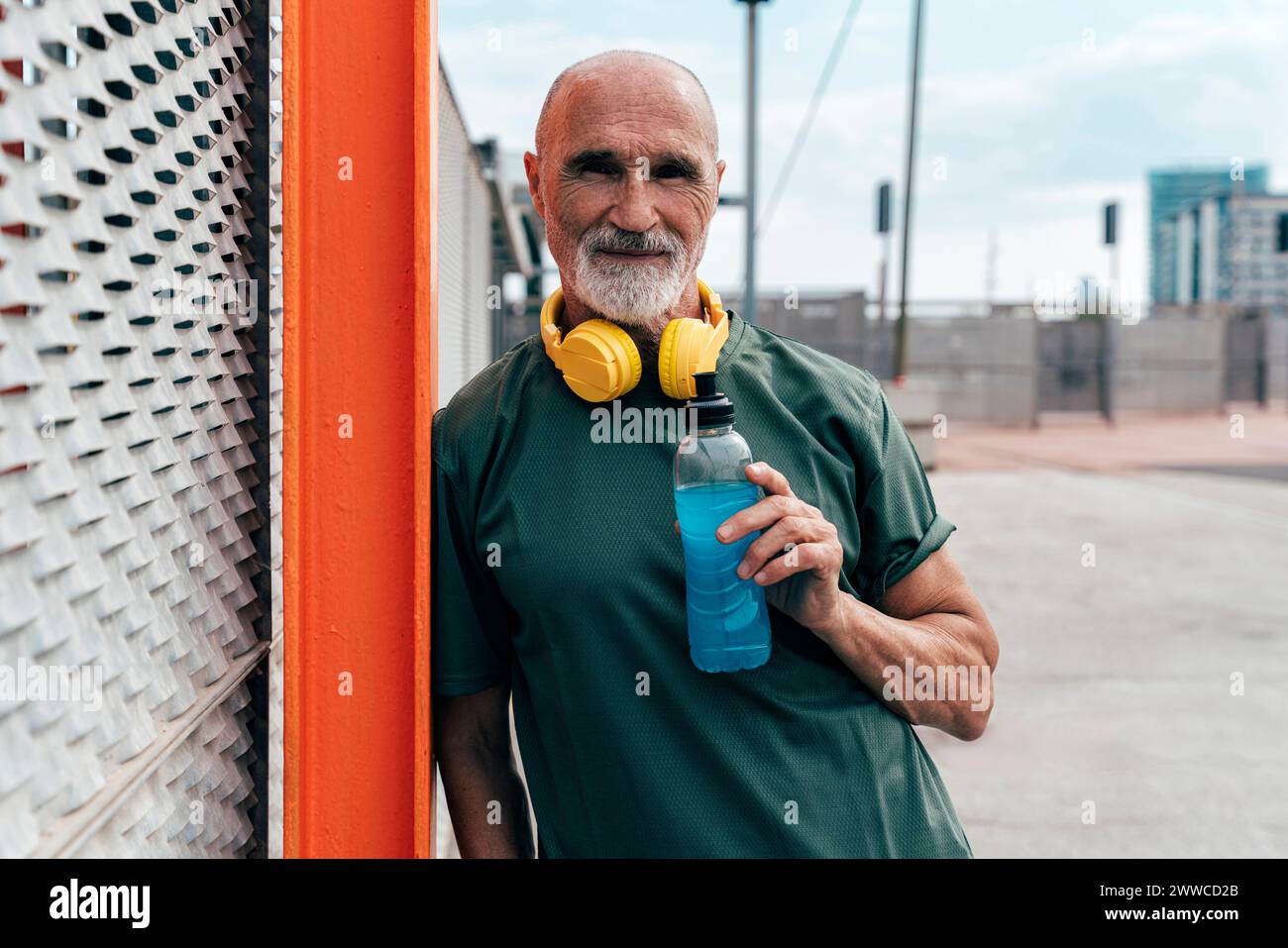 Aktiver Seniorenmann mit kabellosen Kopfhörern, die Energy Drink halten und sich an einen Zaun lehnen Stockfoto