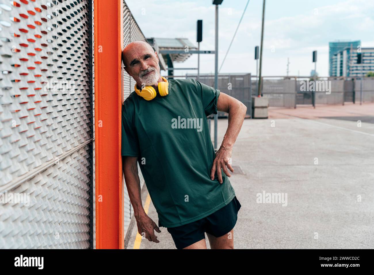 Aktiver Seniorenmann mit kabellosen Kopfhörern, die sich auf einen Zaun stützen Stockfoto