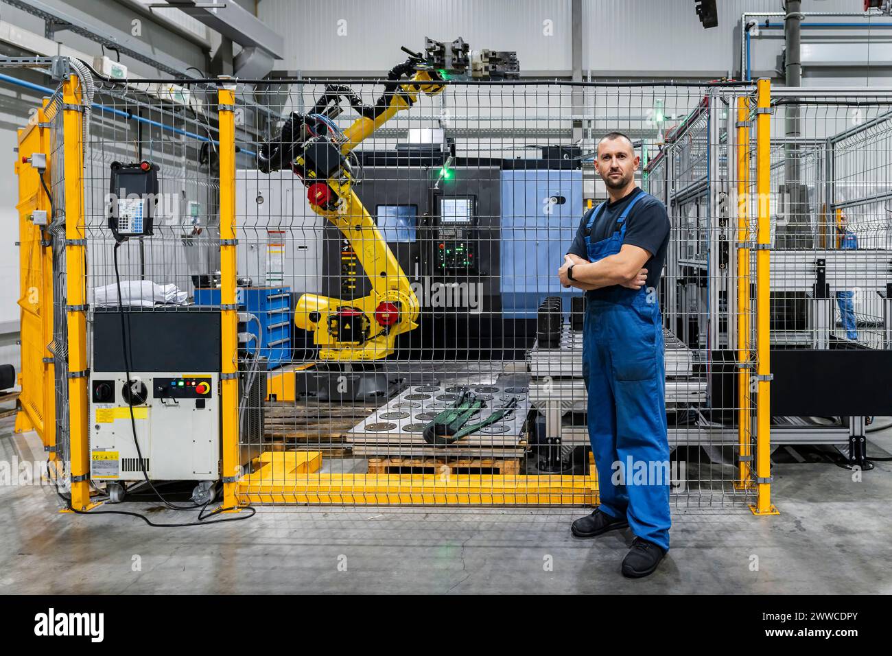 Junger Wartungstechniker, der mit gekreuzten Armen vor dem Roboterarm in einer modernen Fabrik steht Stockfoto