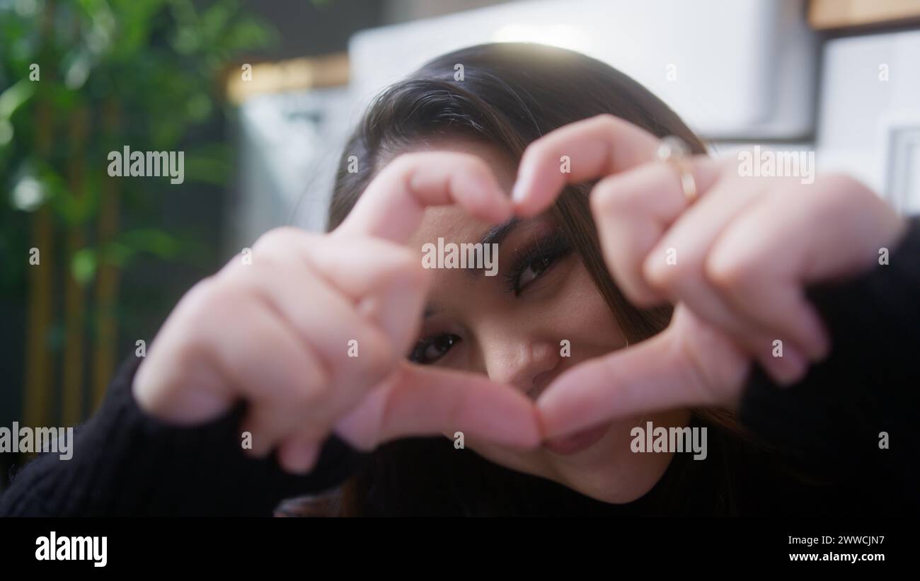 Nahaufnahme eines schönen asiatischen Mädchens, das mit verbundenen Fingern Herzform macht und mit einem Lächeln dazwischen in die Kamera schaut Stockfoto