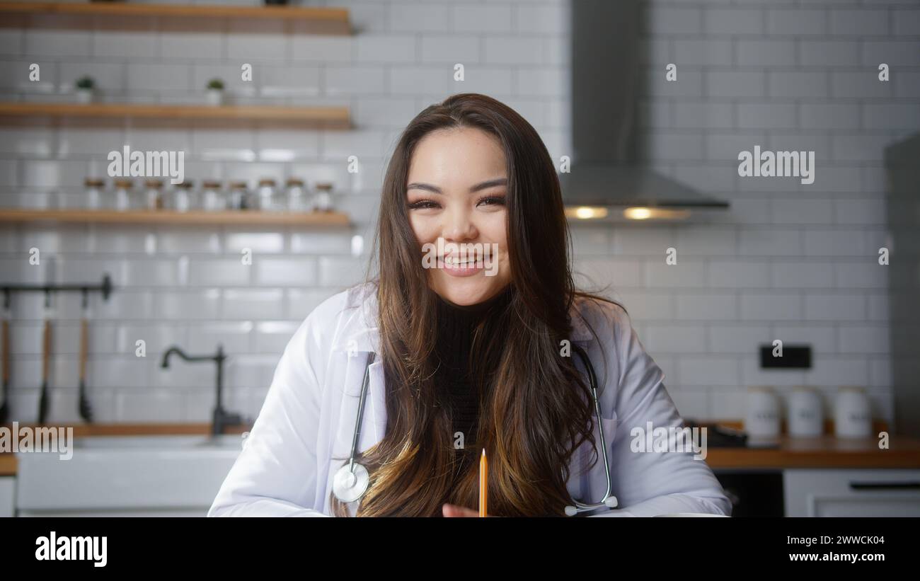 Lächelnde weibliche asiatische Ärztin trägt weiße Uniform mit Stethoskop, die Videokonferenz führt, online mit dem Patienten chatten und die Kamera in die Webcam schauen. Re Stockfoto