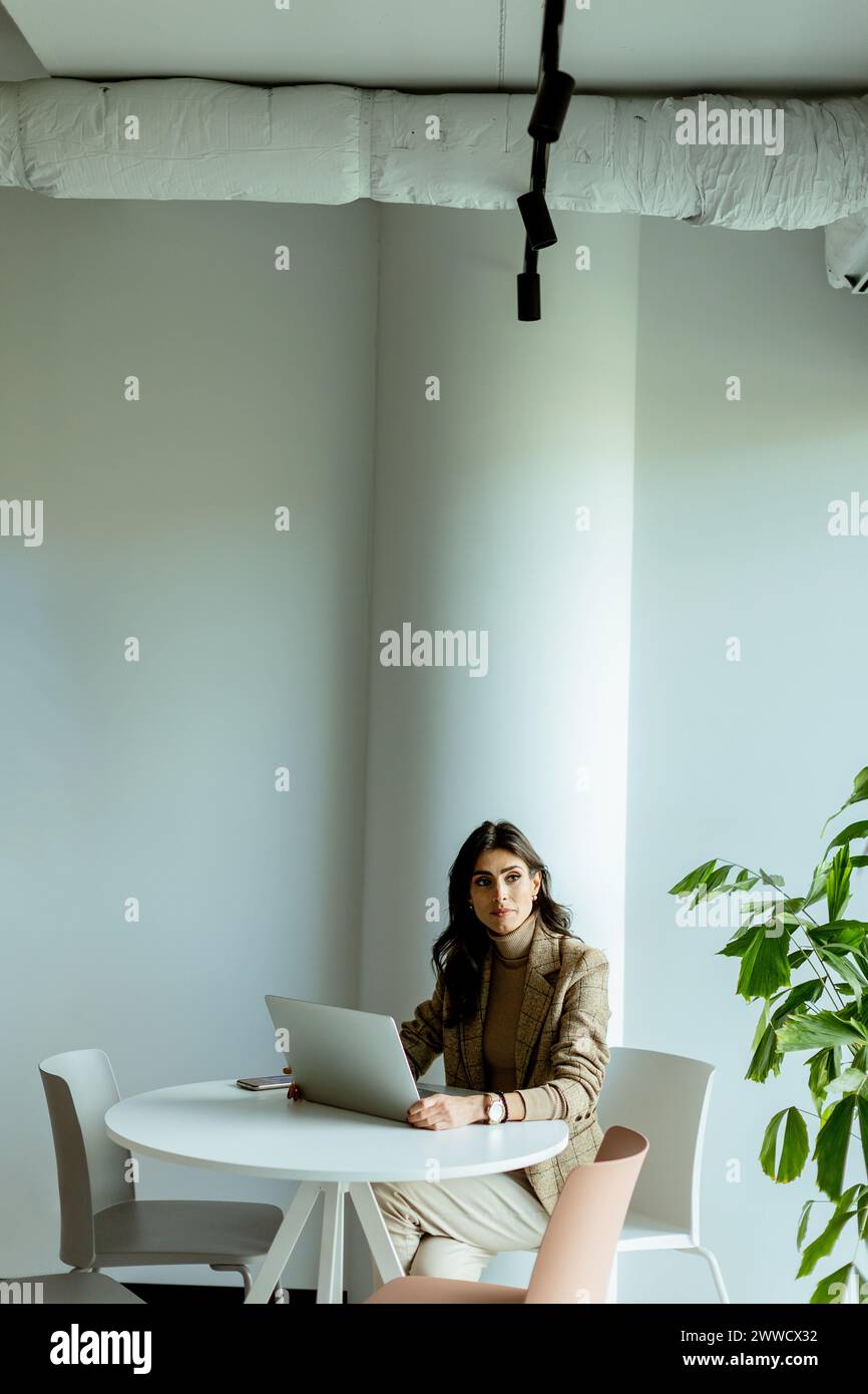 Eine konzentrierte Geschäftsfrau in einem stilvollen Blazer lässt sich auf ihrem Laptop an einem eleganten, weißen Tisch, umgeben von minimalistischem Dekor, verstecken. Stockfoto