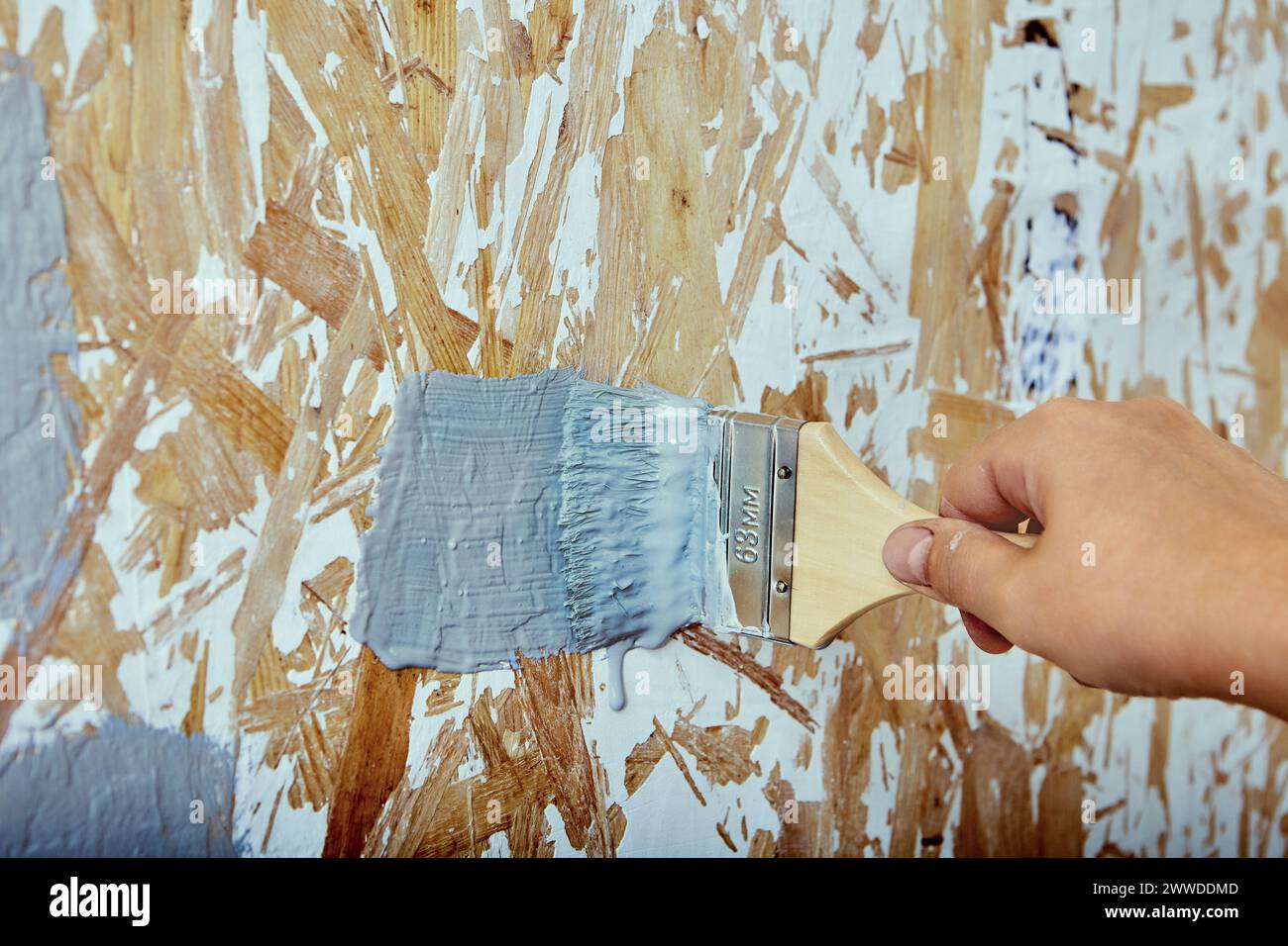 Pinsel in der Hand des Bauarbeiters, Hausmaler wendet Pinselstriche mit grauer Farbe auf die Spanplattenwand an. Stockfoto