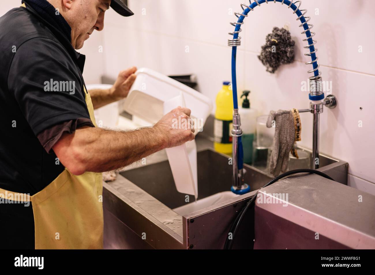 Der Metzger trägt eine Schürze, die im Waschbecken der Metzgerei Waschtische und -Utensilien trägt und im Betrieb Reinigungs- und Wartungsaufgaben durchführt Stockfoto