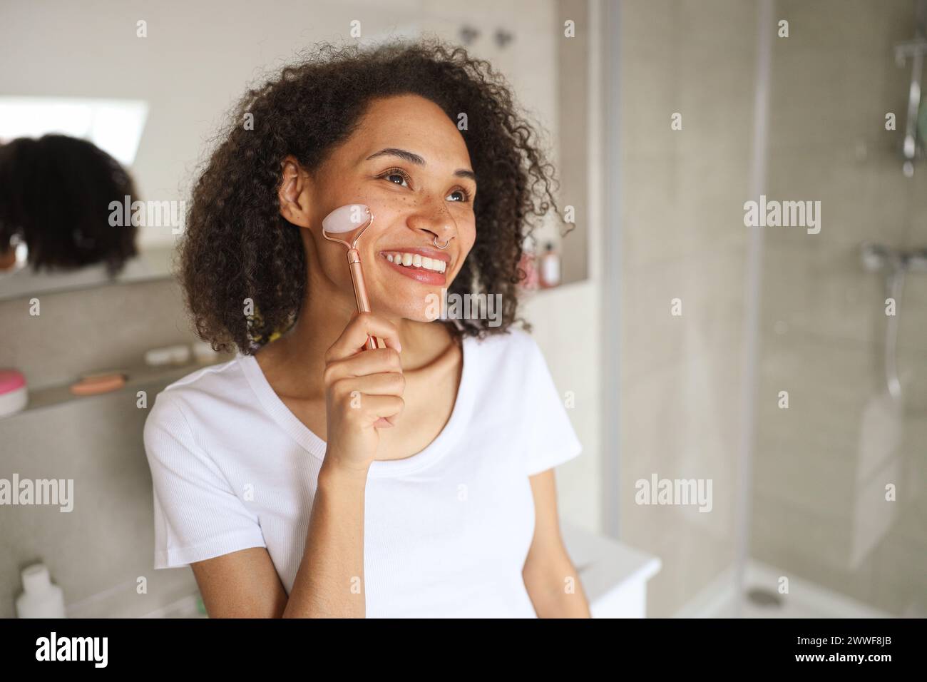 Eine Frau mit schwarzen Haaren lächelt, während sie eine Rosenquarzwalze im Gesicht benutzt Stockfoto