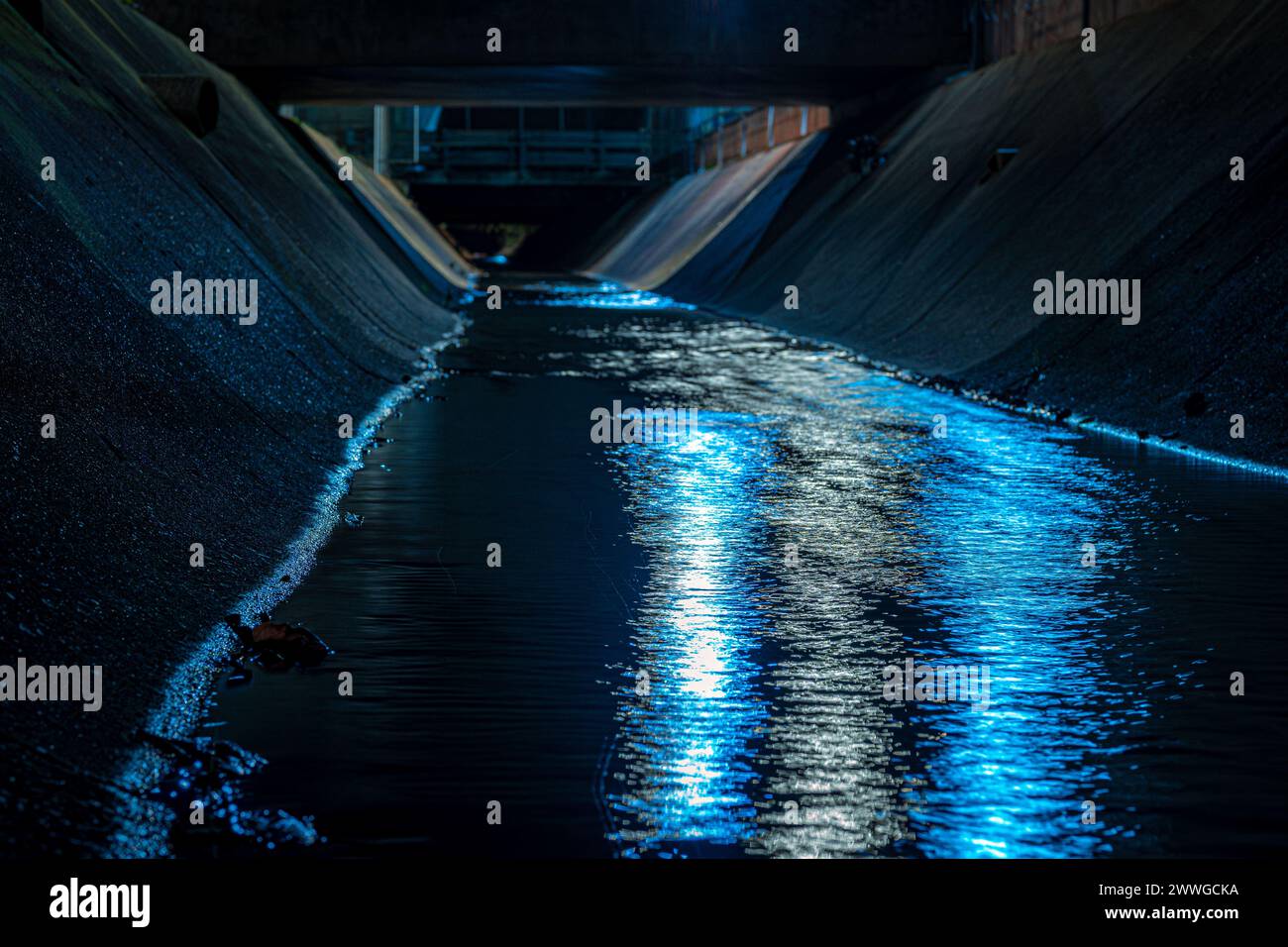 Reflektierender städtischer Wasserweg unter einer Brücke, in blaues Nachtlicht getaucht. Stockfoto Reflektierender städtischer Wasserweg unter einer Brücke, in blaues Nachtlicht getaucht. Stockfoto