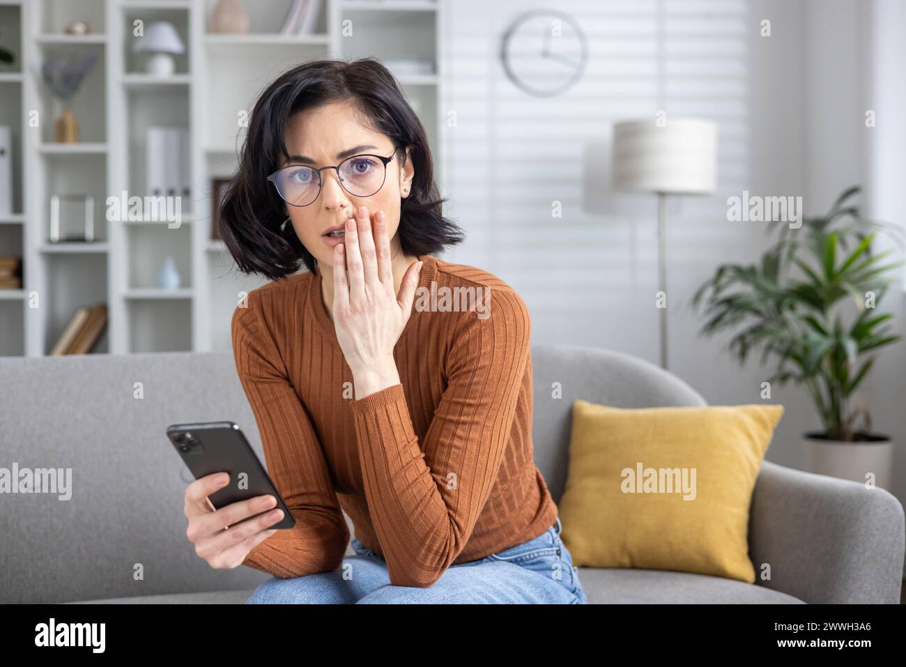 Besorgte Frau, die auf einer Couch sitzt und verwirrt an ihrem Handy aussah, nachdem sie beunruhigende Nachrichten erhalten hatte. Stockfoto
