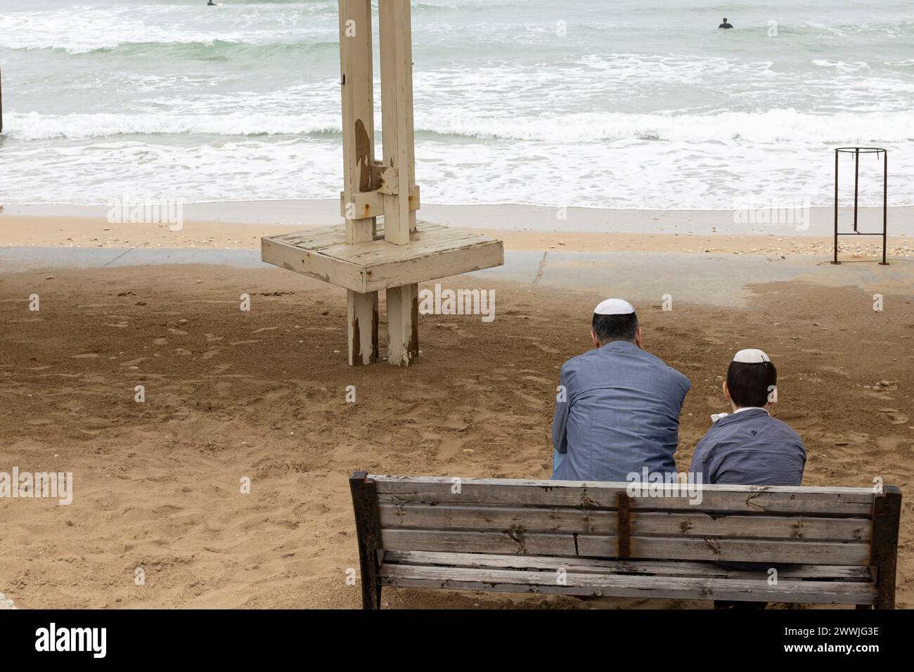 Haifa, Israel - 20. März 2024 sitzen Vater und Sohn in passenden Jacken und Kippahs auf einer Bank am Strand und beobachten die Surfer. Stockfoto