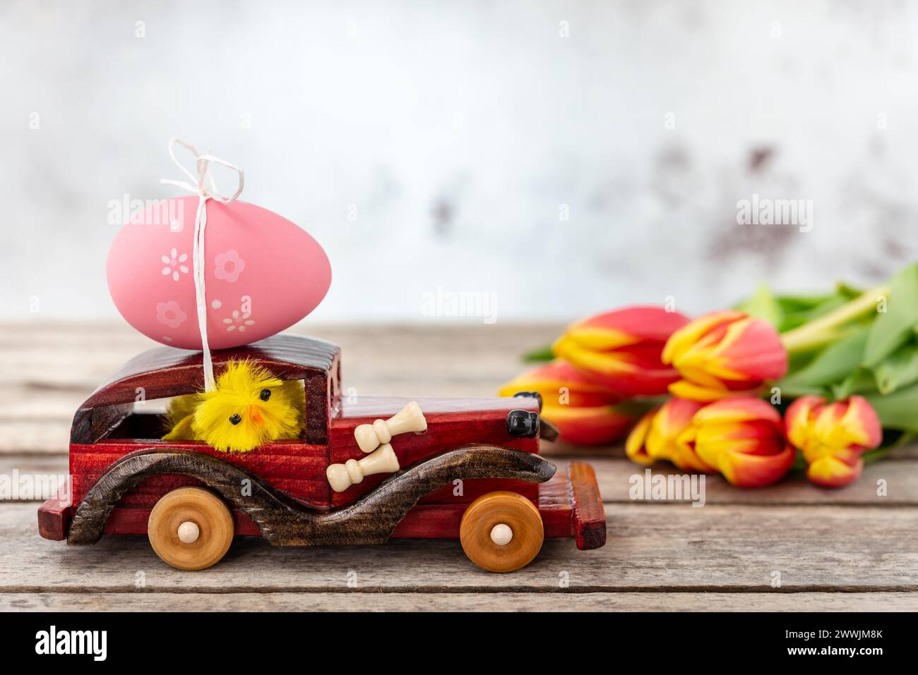 Ein hölzerner Spielzeugwagen mit einem Osterei auf dem Dach und einem Hühnerfahrer neben einem Tulpenstrauß auf einem rustikalen Tisch. Selektiver Fokus. Stockfoto