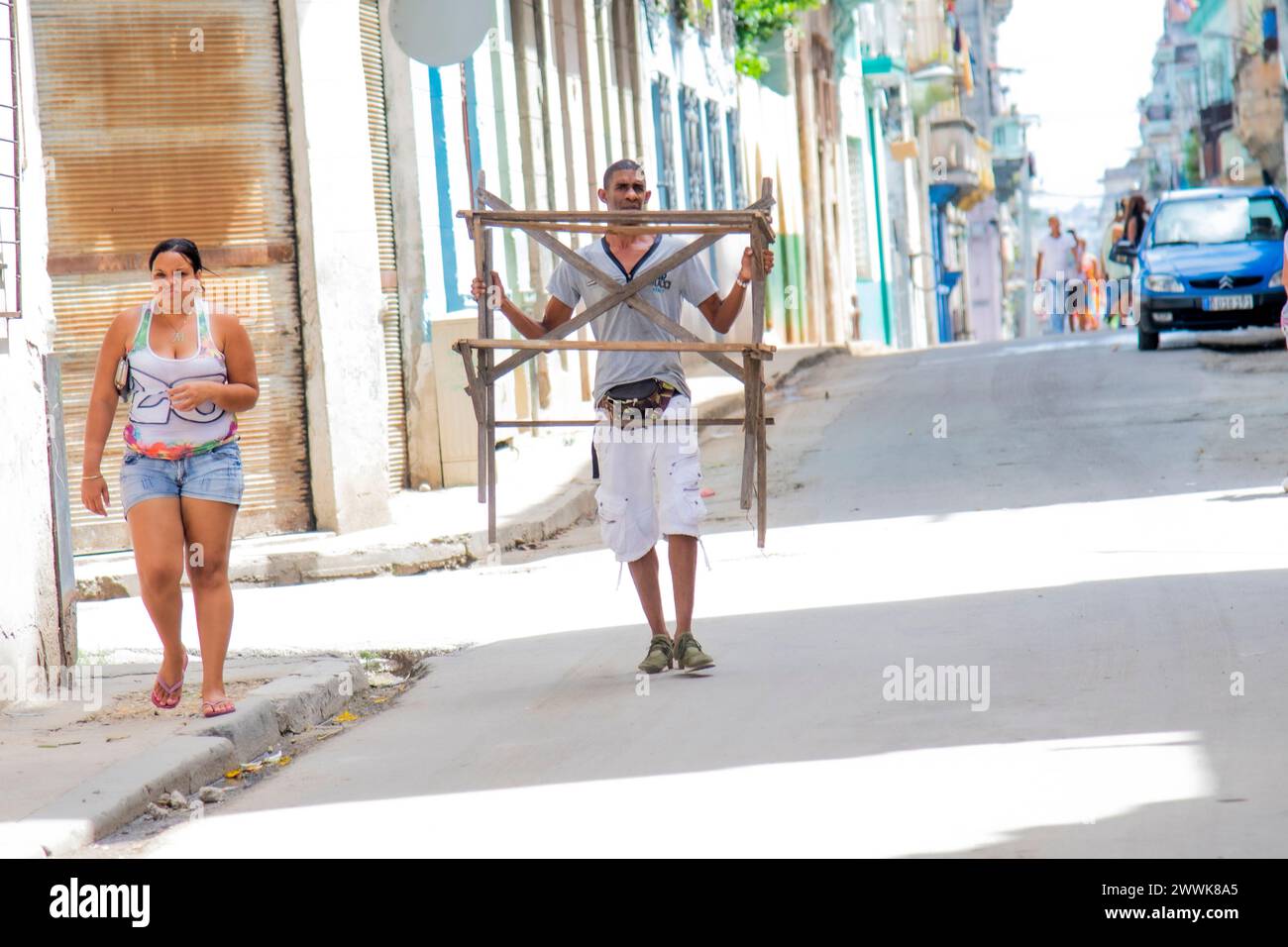 Ein kubanischer Mann trägt ein hölzernes Objekt mitten auf der Straße in Havanna, Kuba, während eine Frau auf dem Bürgersteig entlang läuft. Stockfoto