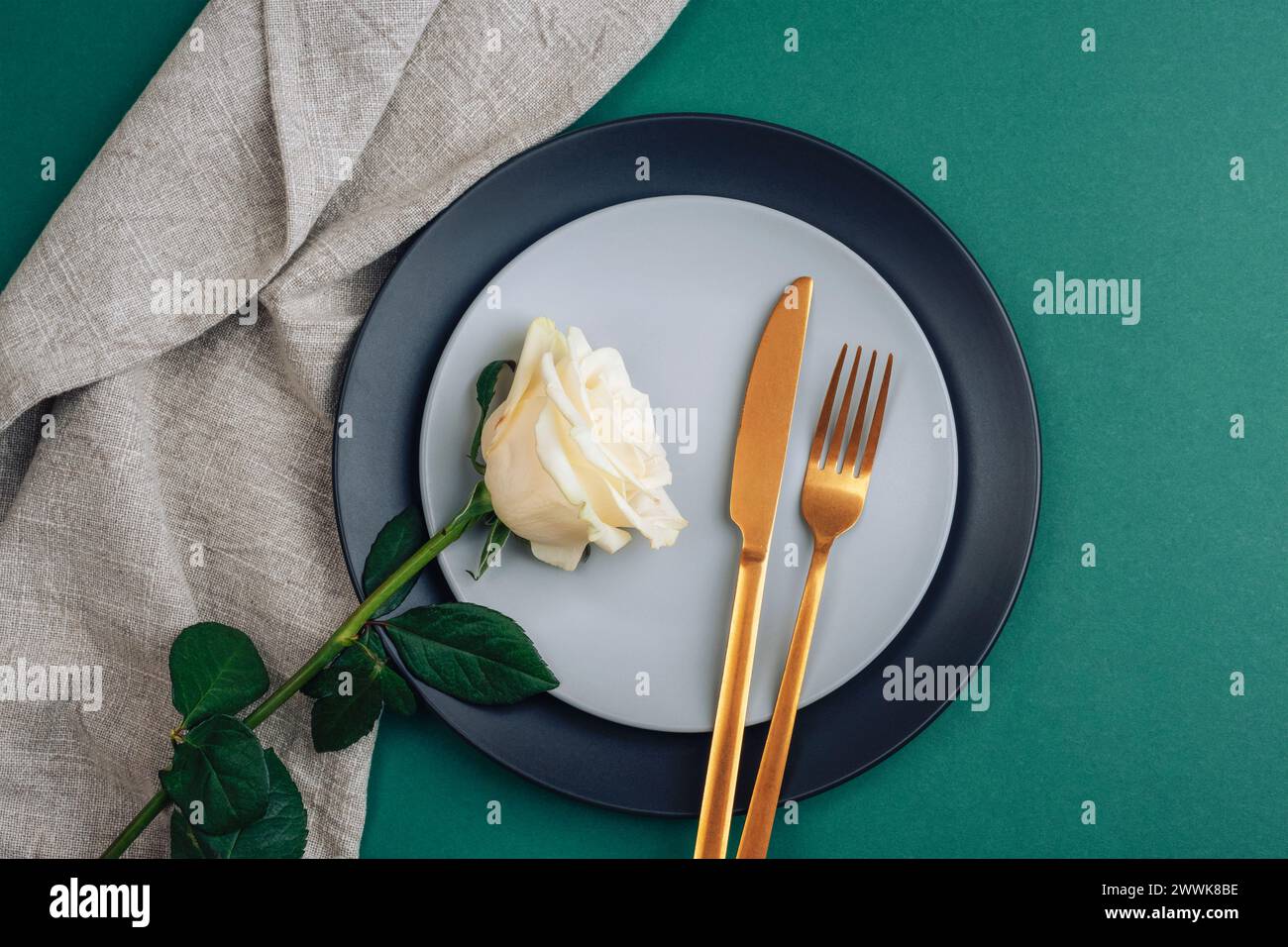 Tischdekoration mit weißer Rosenblüte, Tellern und goldenem Besteck auf dunkelgrünem Hintergrund. Draufsicht, flach. Stockfoto