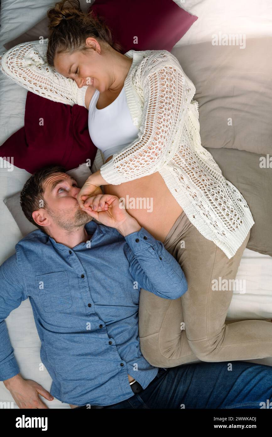 Beschreibung: Werdende Eltern liegen auf dem Sofa, Vater hält die Hand der Mutter sanft und küsst sie. Brillante Aufnahme. Stockfoto