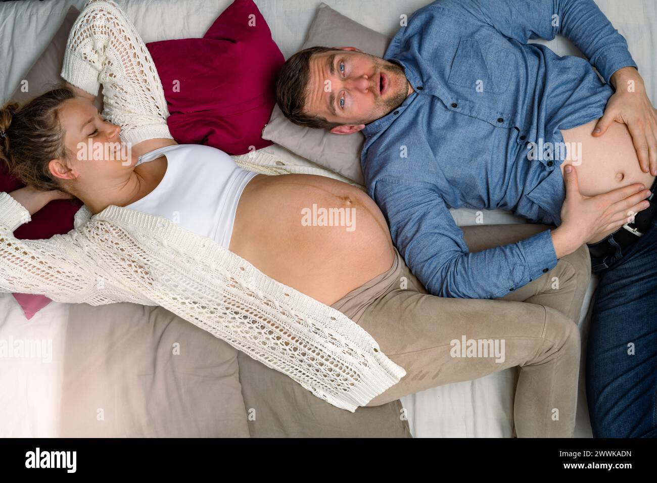 Beschreibung: Werdende Eltern liegen auf dem Sofa und der kopregnante Vater hält den Magen. Brillante Aufnahme. Stockfoto