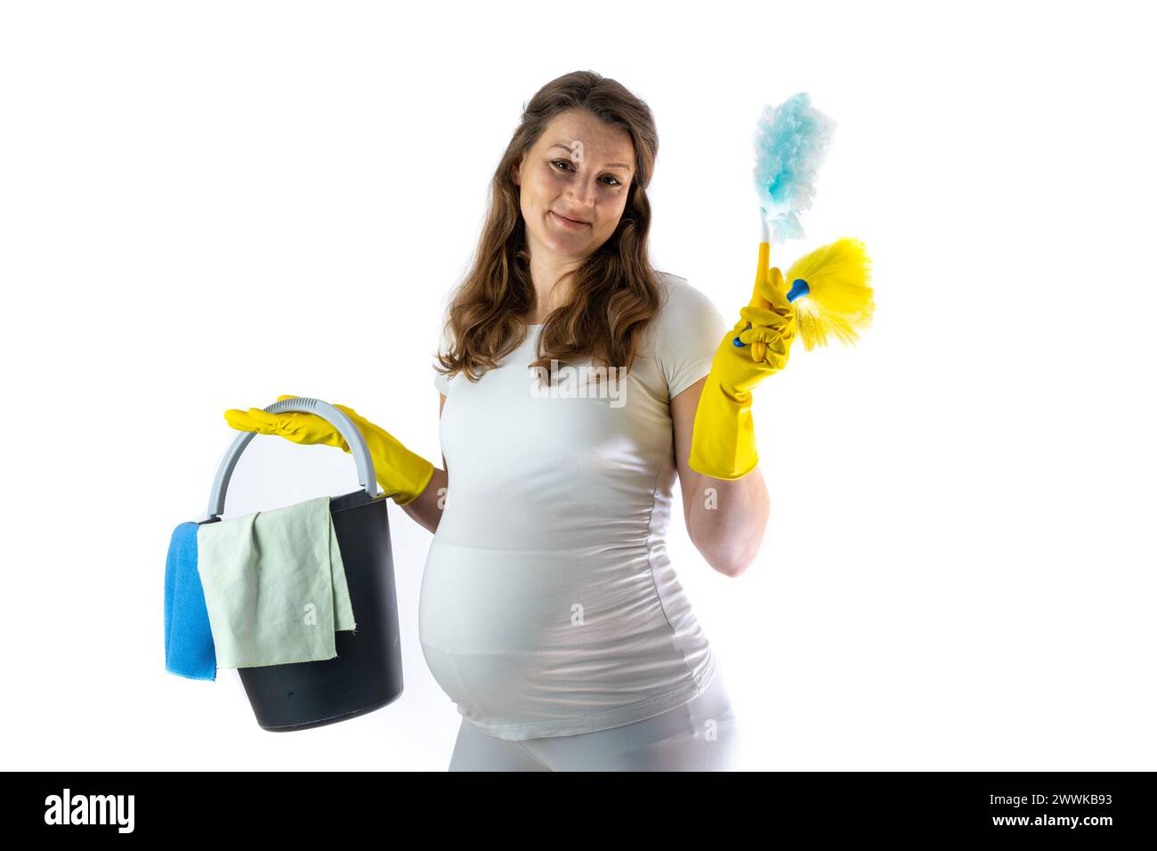 Beschreibung: Schwangere Frau mit rundem Bauch und lächelndem Gesichtsausdruck hält Reinigungsutensilien in den Händen und hat den Drang, die Wohnung zu reinigen. Stockfoto