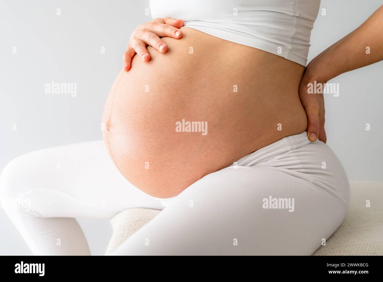 Beschreibung: Mittlerer Teil einer Frau mit stark schwangeren Baby-Unebenheiten, die auf einem Trainingsball sitzen und Rückenschmerzen spüren. Seitenansicht. Weißer Hintergrund. Br Stockfoto