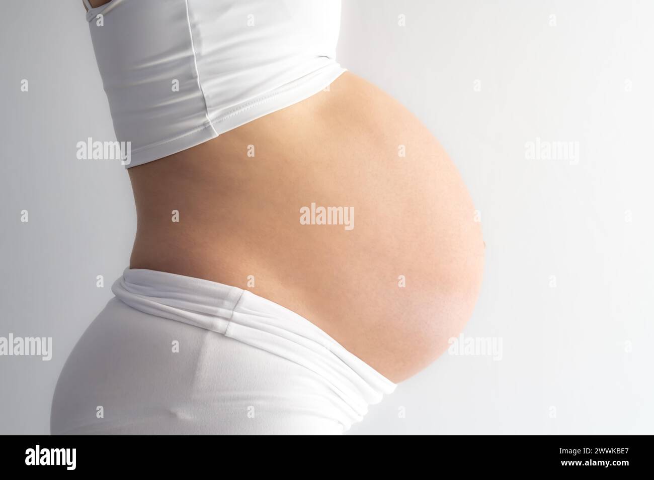 Beschreibung: Mittelteil einer nicht erkennbaren stehenden Frau mit sehr rundem Schwangeren. Seitenansicht. Weißer Hintergrund. Brillante Aufnahme. Stockfoto
