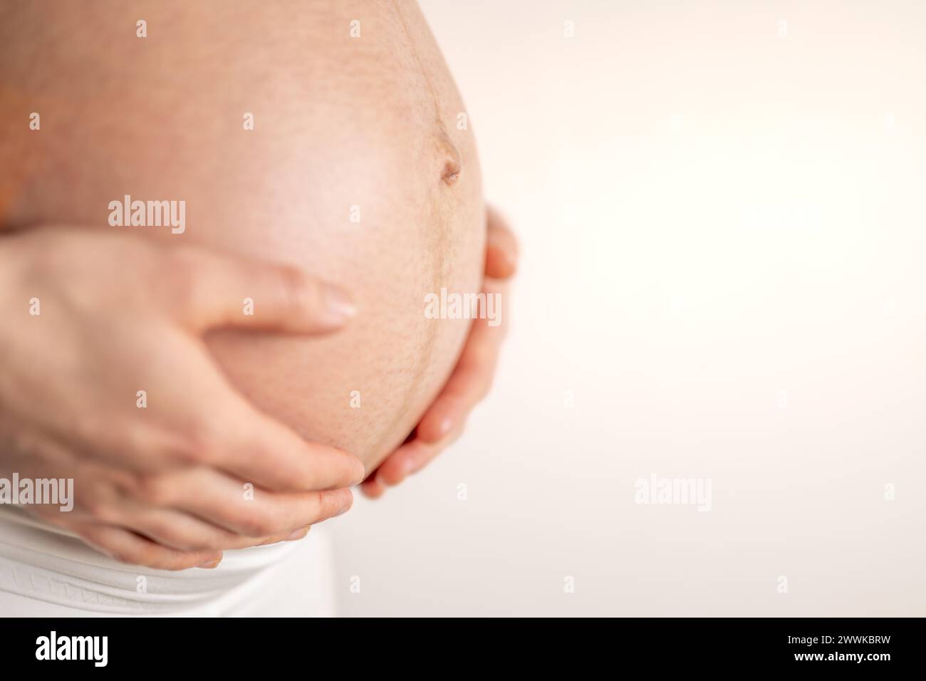 Beschreibung: Schwangere Frau, die im letzten Schwangerschaftsmonat sanft ihren nackten runden Bauch mit den Händen hält. Drittes Trimenon - Woche 36. Nahaufnahme. Seite Stockfoto
