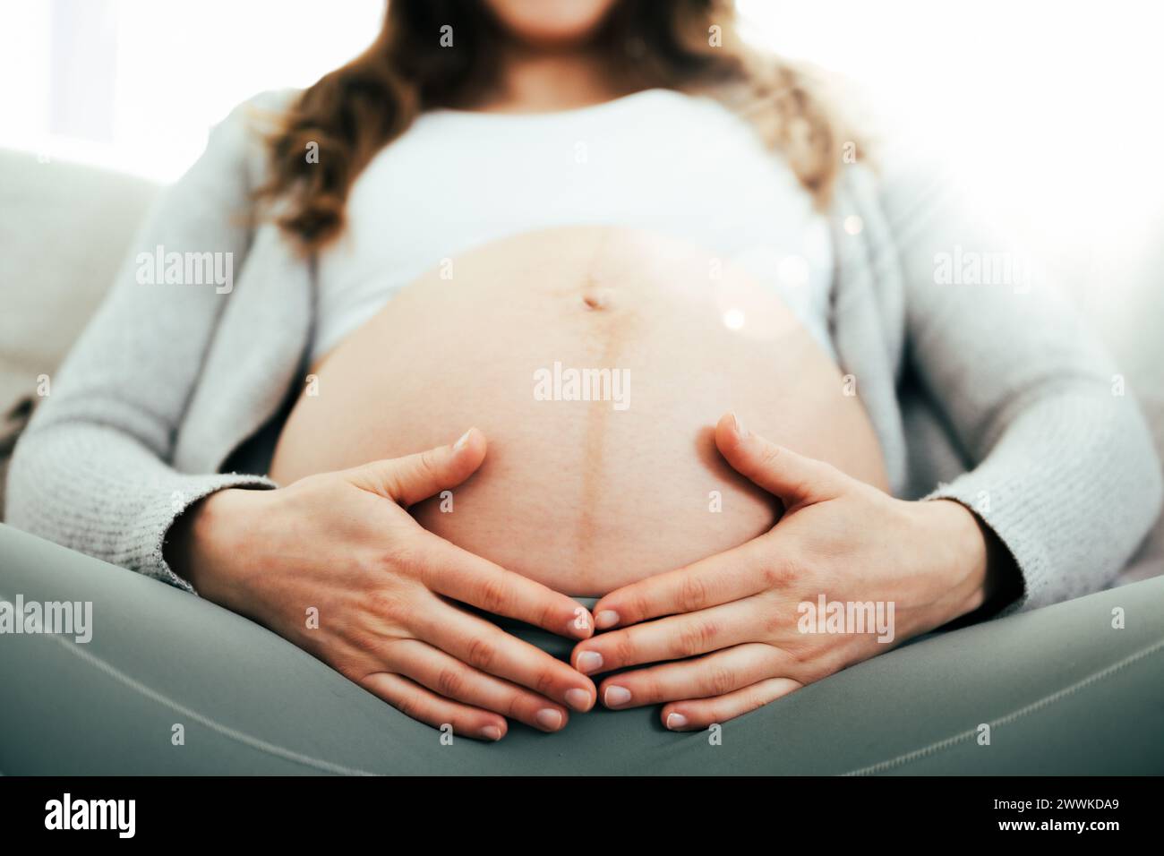 Beschreibung: Frontalansicht einer Frau, die in den letzten Monaten der Schwangerschaft auf dem Sofa sitzt und sanft ihren Bauch hält. Schwangerschaft drittes Trimenon - Woche 34. Frontal Stockfoto