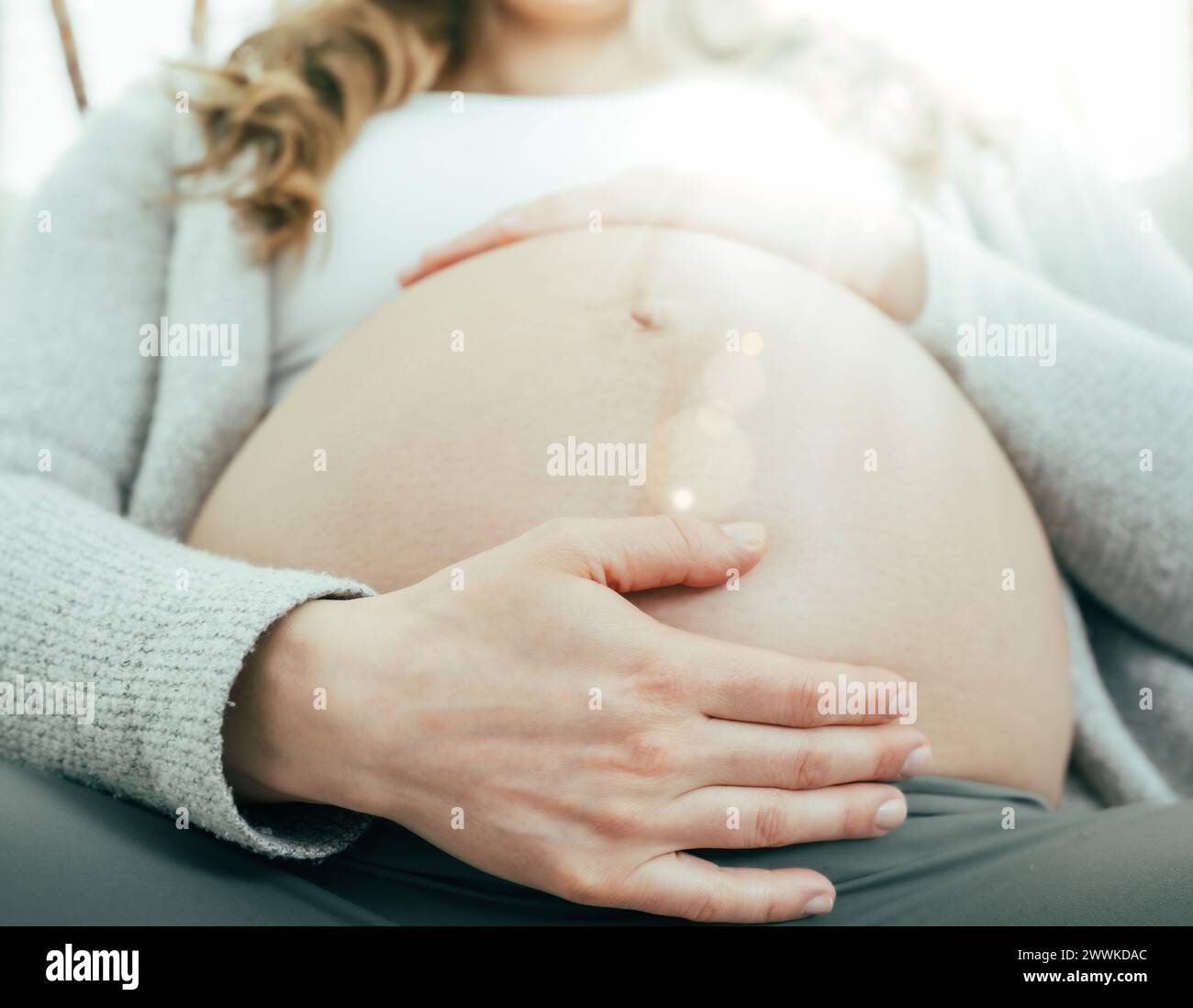 Beschreibung: Frontalansicht einer Frau, die in den letzten Monaten der Schwangerschaft auf dem Sofa sitzt und sanft ihren Bauch hält. Schwangerschaft drittes Trimenon - Woche 34. Frontal Stockfoto