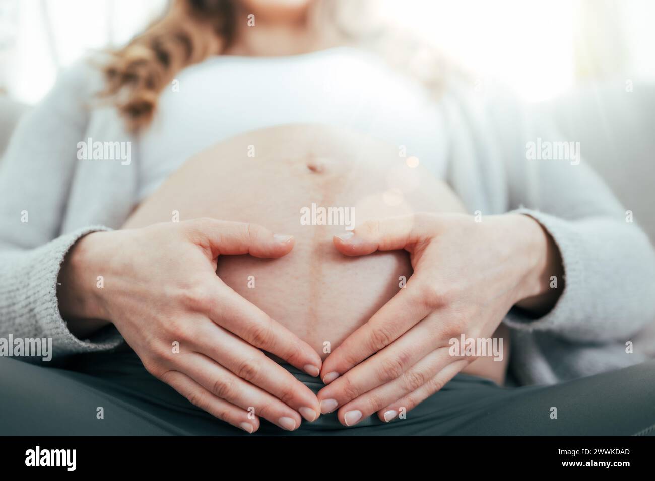 Beschreibung: Niedriger Blickwinkel einer Frau, die in den letzten Monaten der Schwangerschaft auf dem Sofa sitzt und sanft ihren Bauch hält. Schwangerschaft drittes Trimenon - Woche 34. Vorne Stockfoto