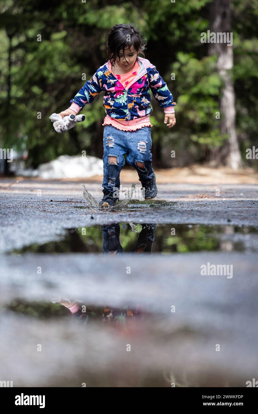 Ein junges Mädchen spielt im Regen und plätschert in Pfützen. Die Szene ist verspielt und fröhlich. Das Mädchen genießt den Moment und die feuchte Umgebung Stockfoto