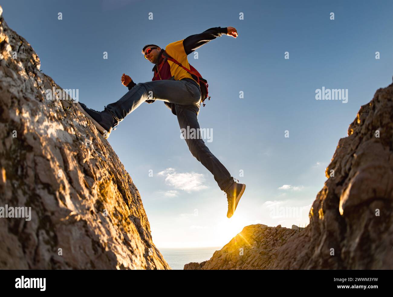 Junger sportlicher Wanderer mit Rucksack springt gegen den Sonnenuntergangshimmel auf den großen Felsen. Aktives Lifestyle-Konzept Stockfoto