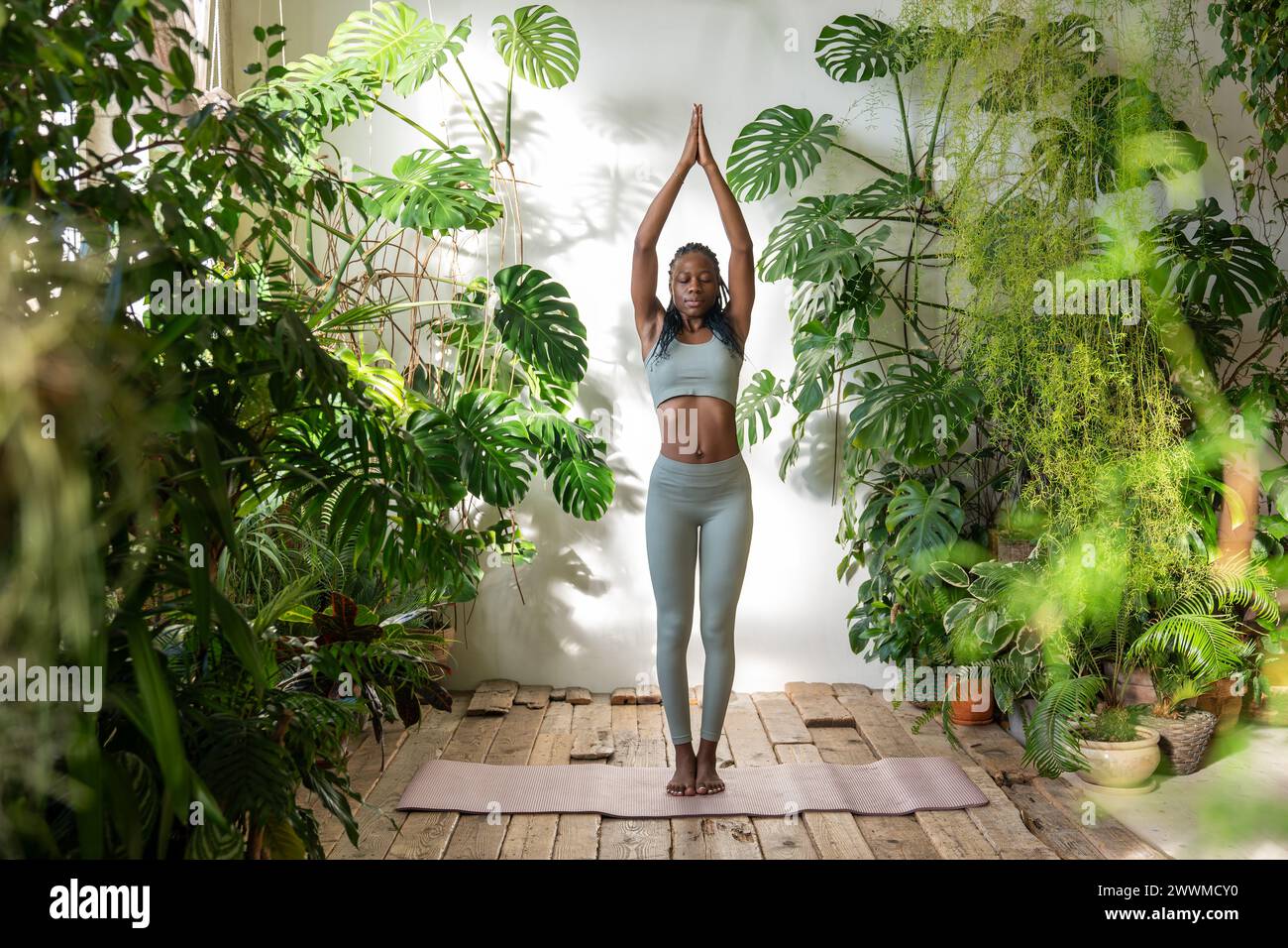 Eine ruhige, ausgewogene schwarze Frau steht auf einer Yogamatte umgeben von Pflanzen und meditiert Ruhe, Harmonie Stockfoto