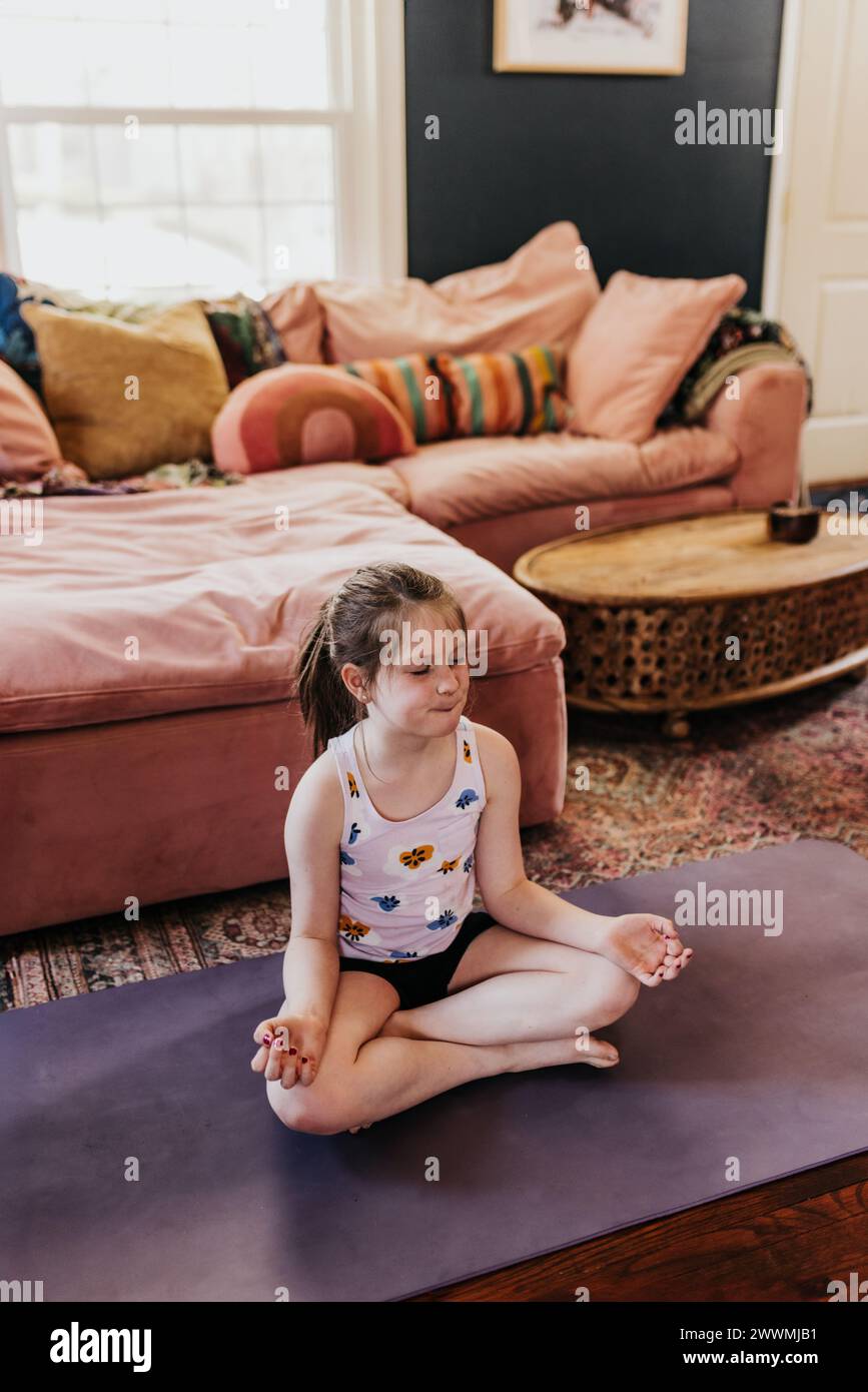 Das junge Mädchen meditiert, während es im Wohnzimmer auf einer violetten Yogamatte sitzt Stockfoto