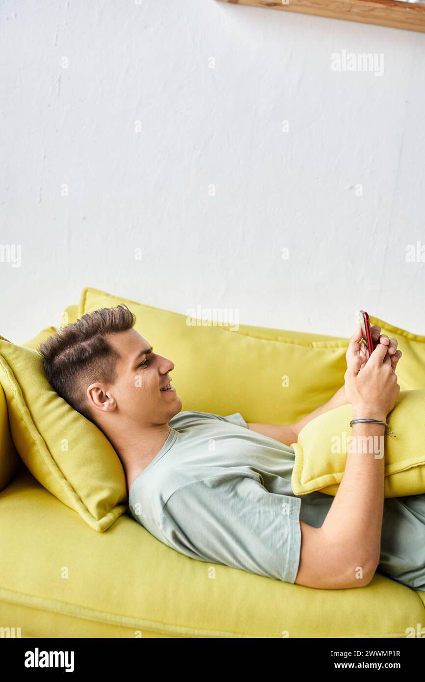 Profil eines fröhlichen Schülers, der im Wohnzimmer auf der gelben Couch liegt und in soziale Medien scrollt Stockfoto
