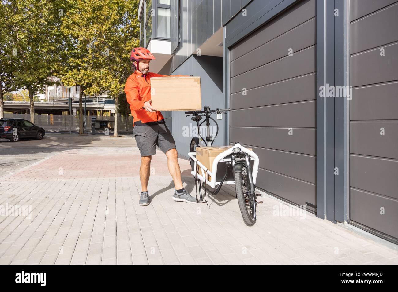 Junger Kurier mit roter Kleidung und Helm, der auf einem Lastenrad unterwegs ist, kommt am Versandort an, um ein Paket an eine Adresse in der Stadt zu liefern Stockfoto