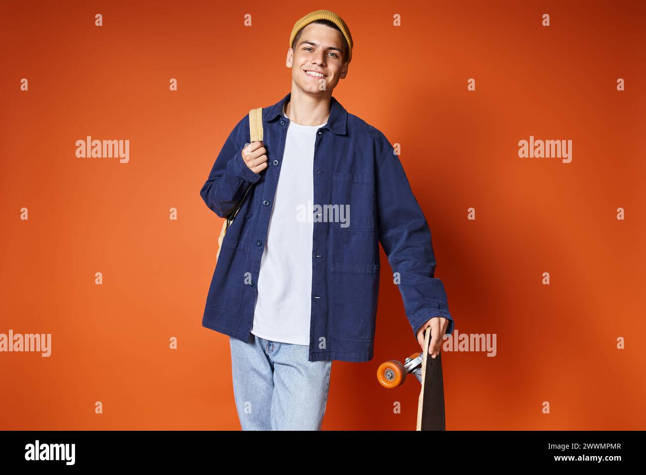 Charismatischer junger Mann im lässigen Outfit, der mit Skateboard vor Terrakotta-Hintergrund steht Stockfoto