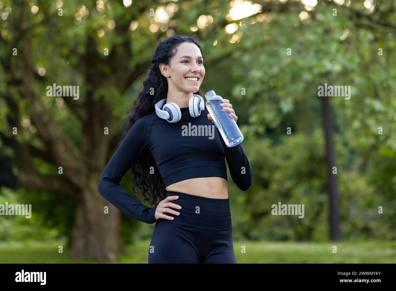 Lächelnde aktive Frau in Trainingsausrüstung, die eine Pause mit einer Wasserflasche in einem grünen Park macht, Kopfhörer um den Hals. Stockfoto