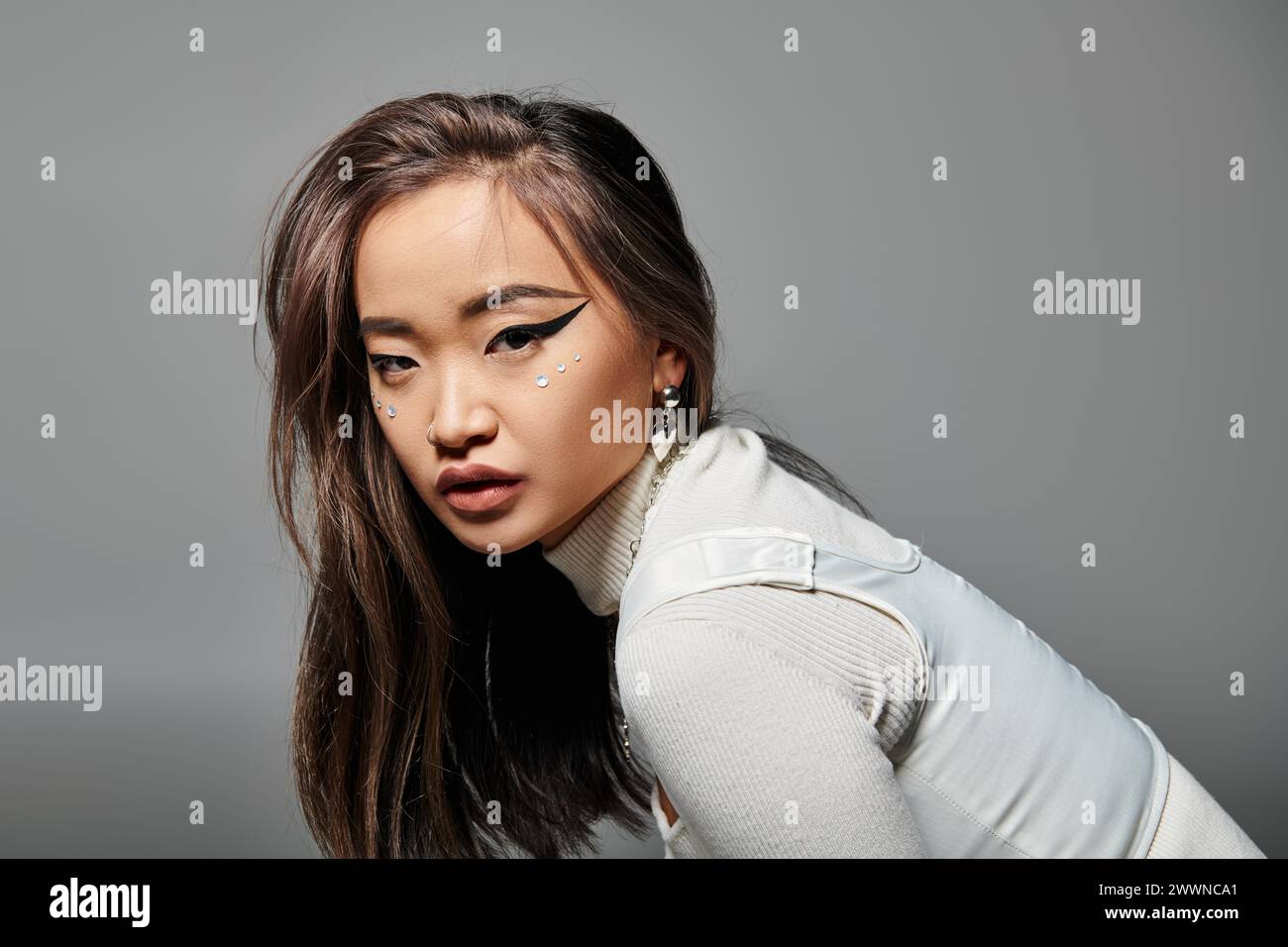 Hübsche asiatische Frau in ihren 20ern mit schwerem Make-up seitlich nach vorne geneigt auf grauem Hintergrund Stockfoto