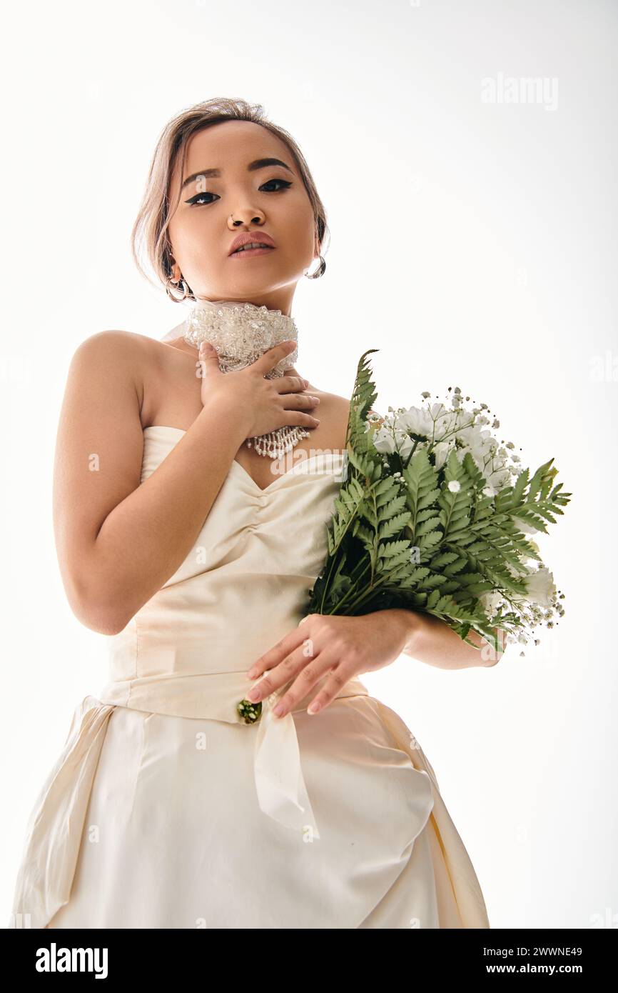 Verführerische junge asiatische Braut in weißer Halskette, die mit Blumenstrauß in die Kamera schaut Stockfoto