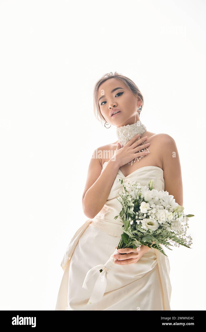Attraktive asiatische Frau berührt weiße Halskette und hält Blumenstrauß auf hellem Hintergrund Stockfoto
