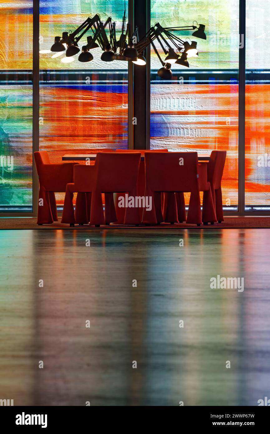 Tisch und Stühle im Bürobereich, vor mehrfarbiger Glasfassade, mit Reflexion auf dem Boden und Deckenlampe im Spinnenstil. Niederländisches Institut Für Sound Stockfoto