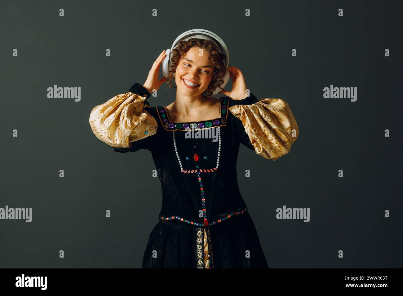 Porträt einer lächelnden jungen Aristokratin mit Kopfhörern und berührendem Kopf und in einem mittelalterlichen Kleid gekleidet lauscht Musik Stockfoto