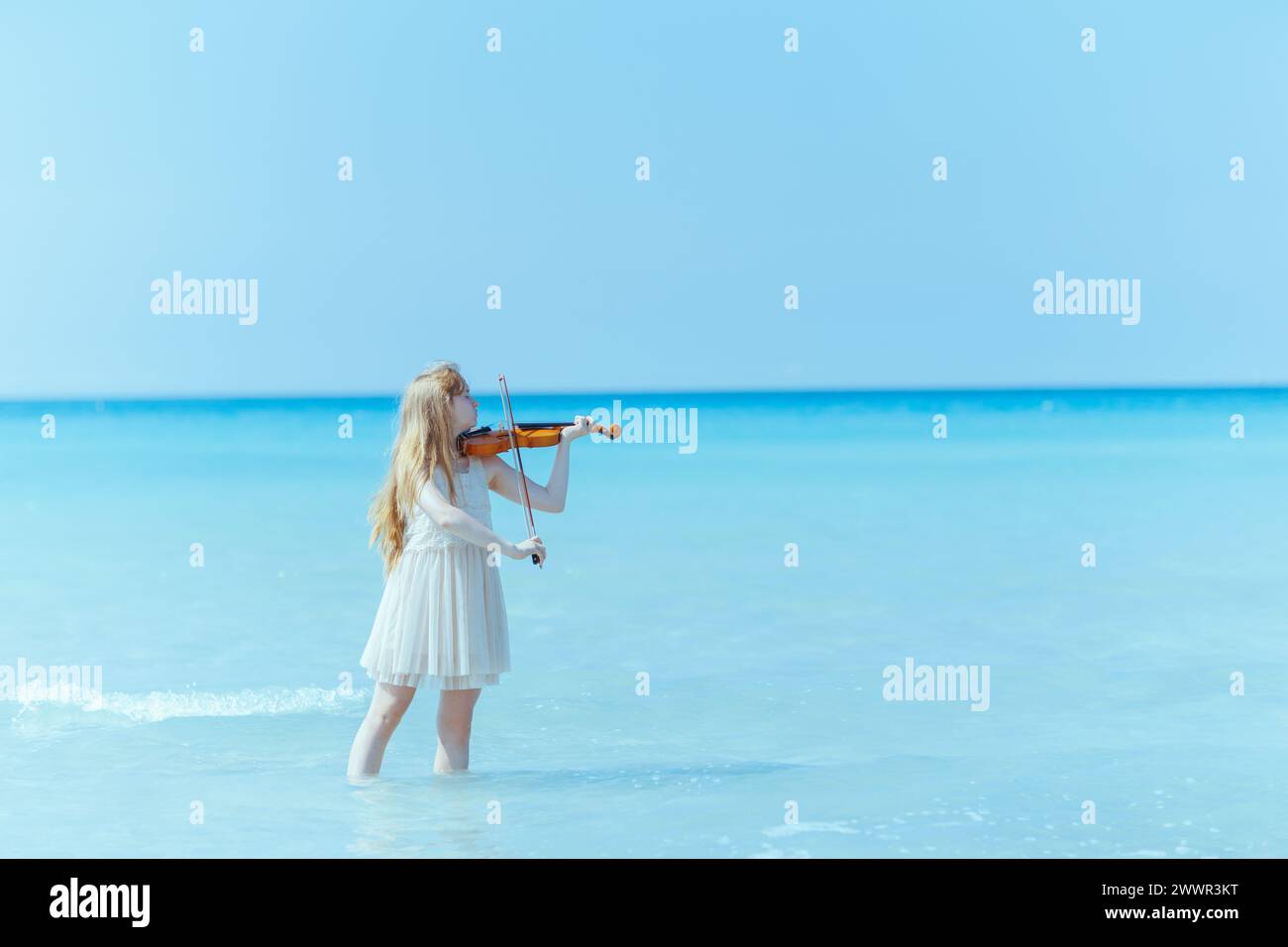 Modernes Mädchen in weißem Kleid an der Meeresküste mit Geige, die gerne spielen. Stockfoto