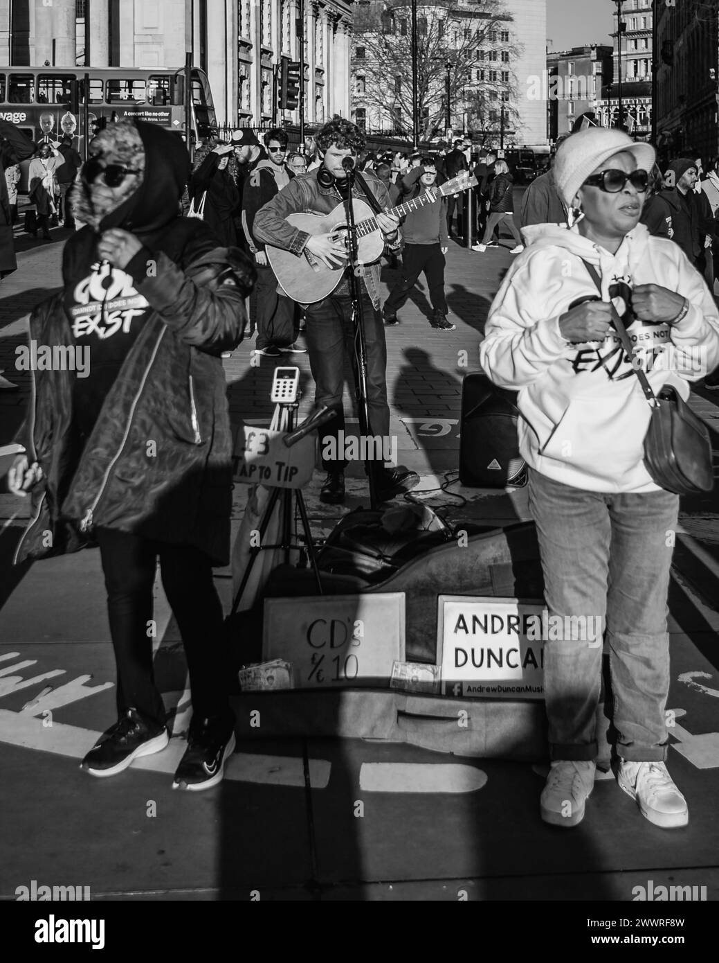 Ein Schwarzweiß-Bild eines Straßenhändlers, der geduldig wartet und sich auf die Bühne stellt, während die Streams of Joy-Gruppe einen spontanen christlichen Sing-A-Long in London dirigiert. Stockfoto