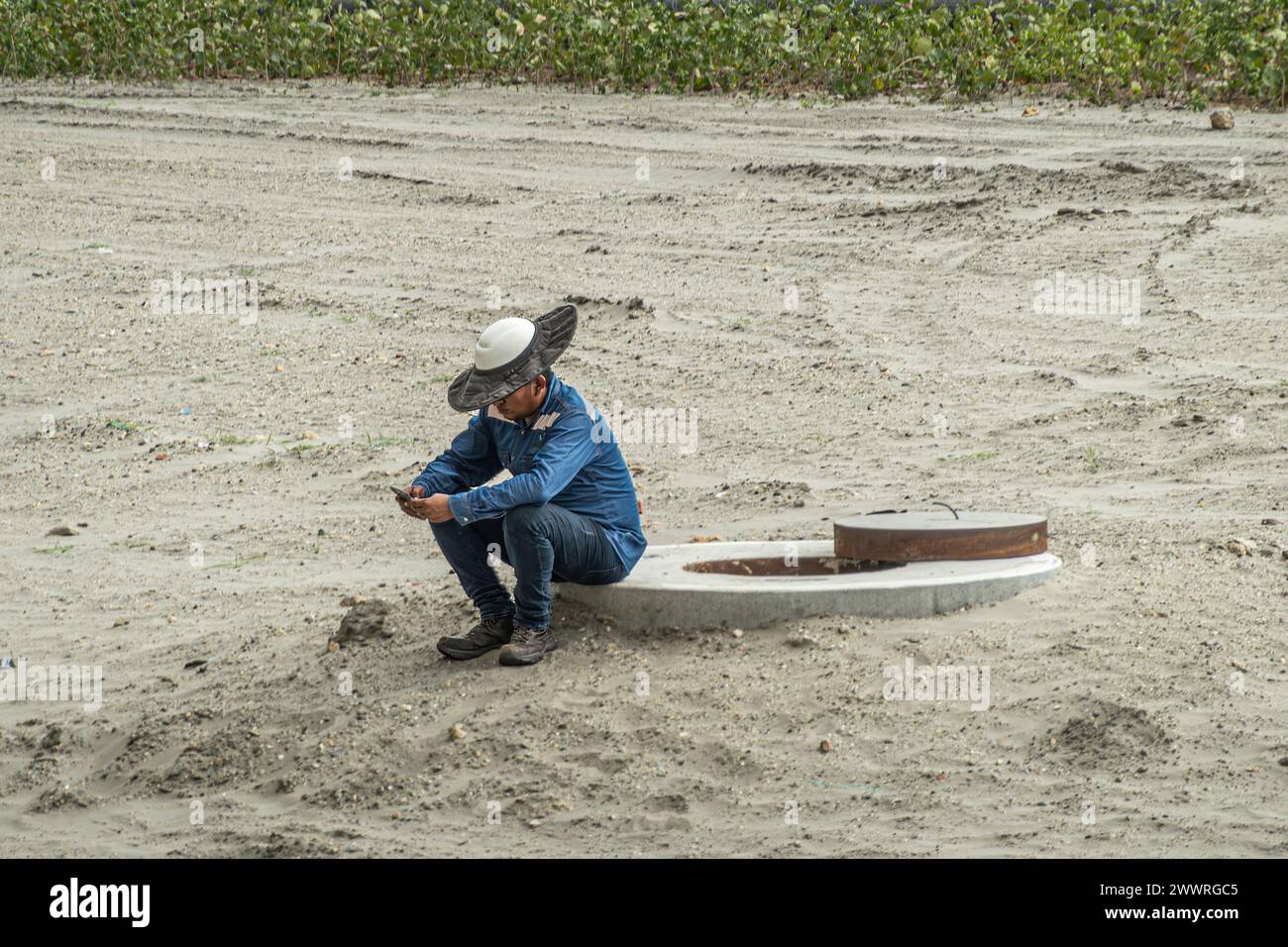 Cartagena, Kolumbien - 25. Juli 2023: Nahaufnahme eines Arbeiters mit Helm und kreisförmiger Sonnencreme sitzt auf Sand und macht eine Pause, um das Smartphone zu überprüfen Stockfoto