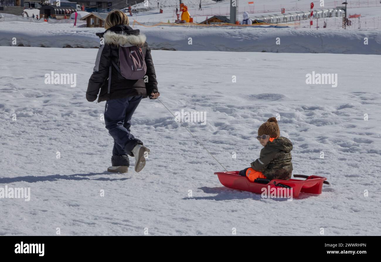 Ein Kleinkind, das auf einem Plastikschlitten sitzt, wird von einer Frau über den gefrorenen See im französischen Skigebiet Tignes gezogen. Stockfoto