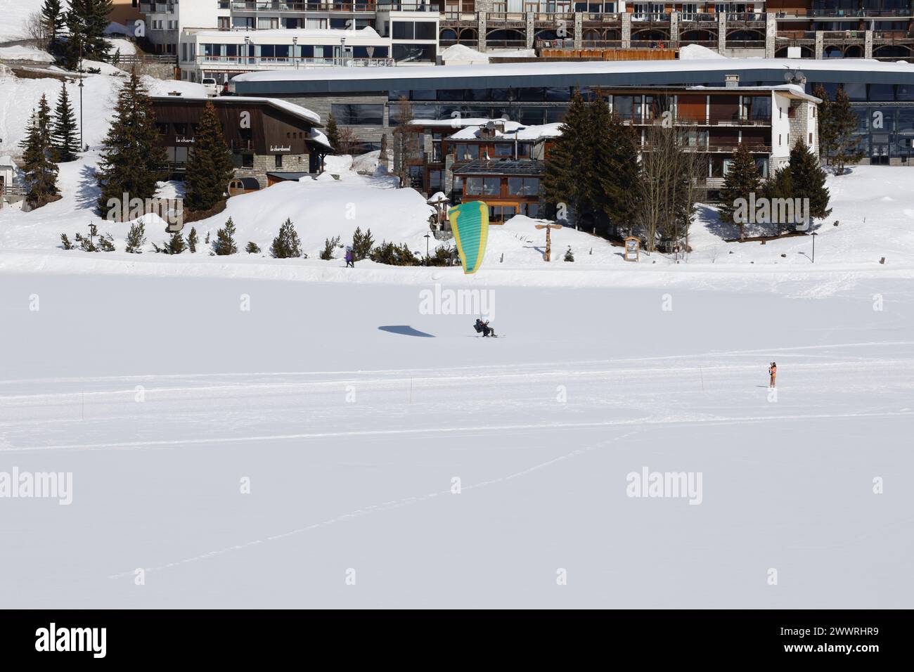 Ein Gleitschirm-Tandem landet vor Bauwerken auf dem schneebedeckten, gefrorenen, natürlichen See in Tignes, einem hochgelegenen Skigebiet in den französischen Alpen. Stockfoto