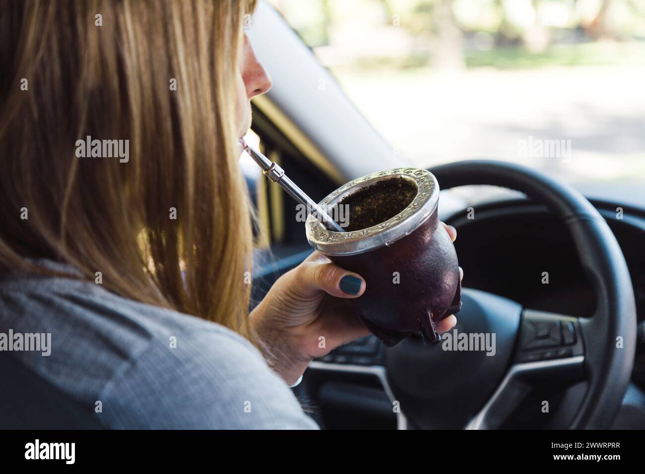 Blonde Frau mittleren Alters, die in einem Auto sitzt und Yerba-Mate-Infusion in einem Mate-Becher trinkt. Stockfoto