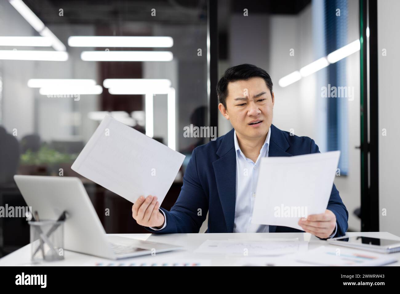 Ein knackiger Mann, der Dokumente hält, während er am weißen Schreibtisch im Büro sitzt. Gutaussehender asiatischer Mann, der auf dem Hintergrund auf der Glaswand des Business Centers arbeitet. Konzept von Technologie und Mensch. Stockfoto