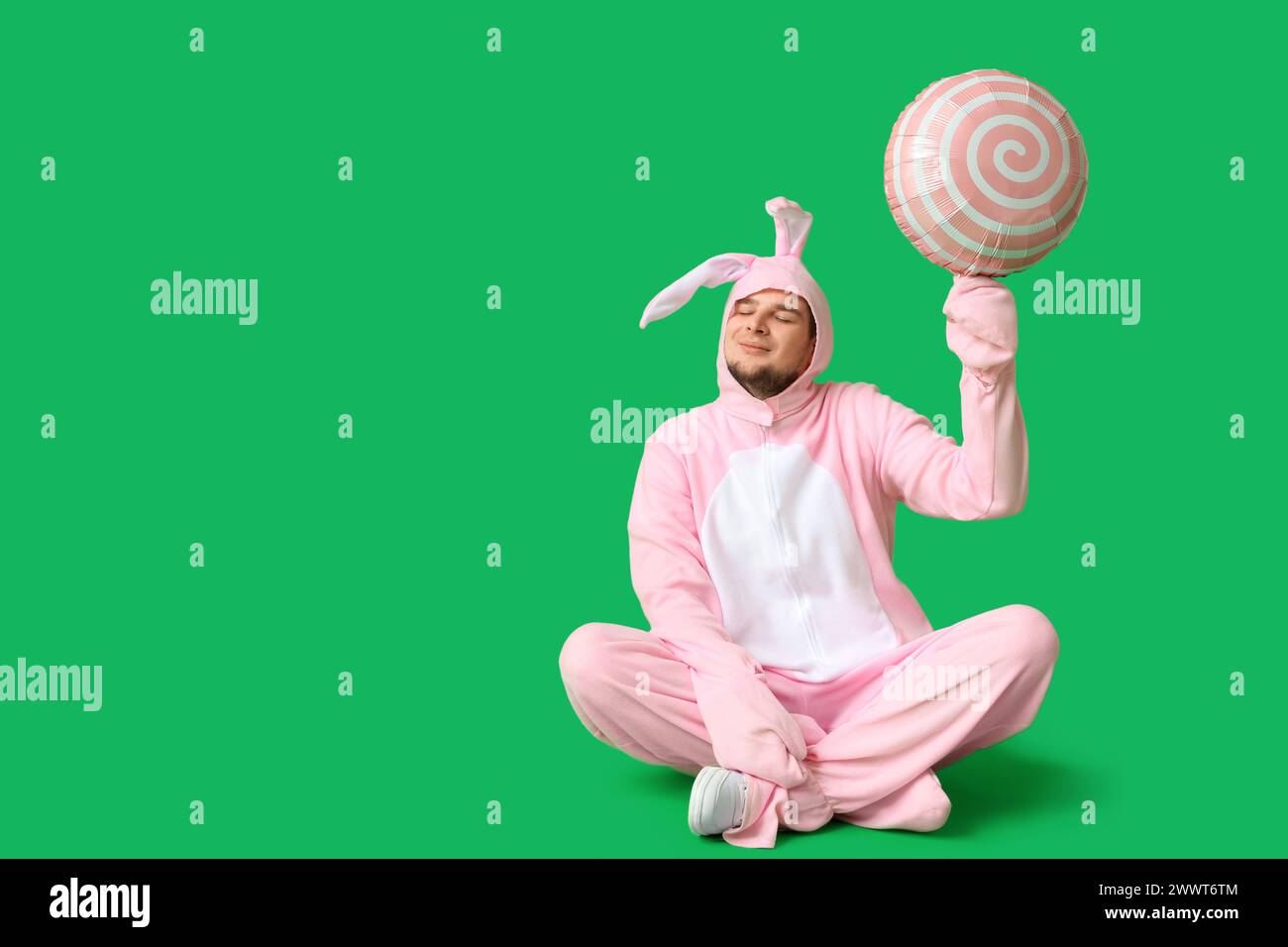 Junger Mann im Hasenkostüm mit Luftballon in Form eines Lutschers, der auf grünem Hintergrund sitzt. Osterfeier Stockfoto