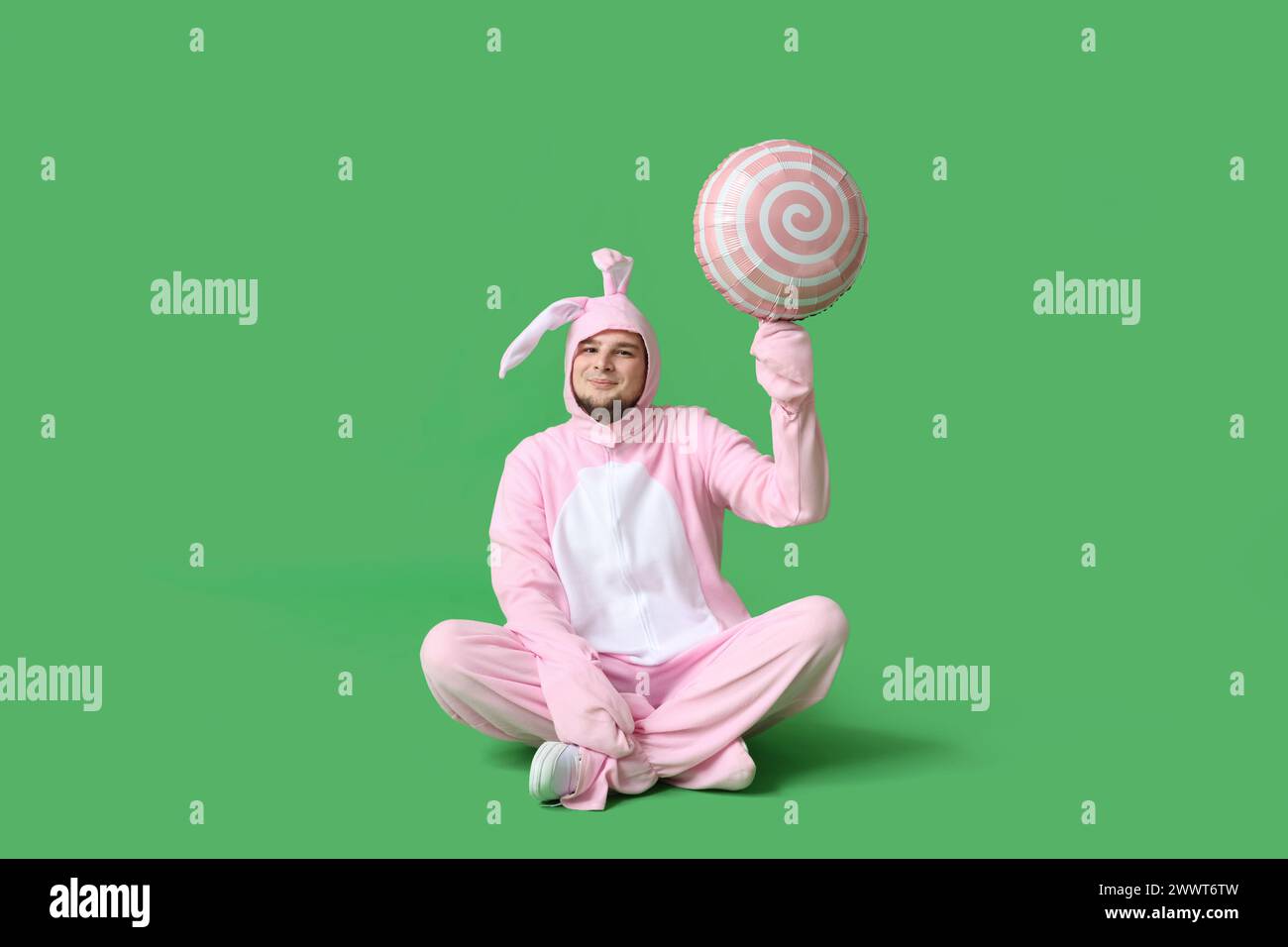 Junger Mann im Hasenkostüm mit Luftballon in Form eines Lutschers, der auf grünem Hintergrund sitzt. Osterfeier Stockfoto