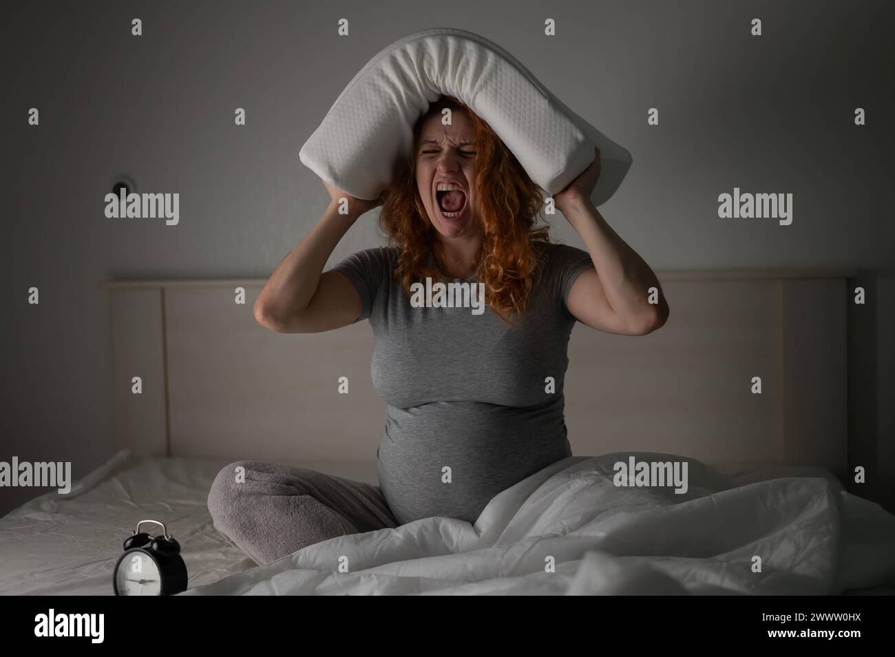 Eine schwangere Frau sitzt im Bett, hält ein Kissen auf dem Kopf und schreit. Schlaflosigkeit. Stockfoto