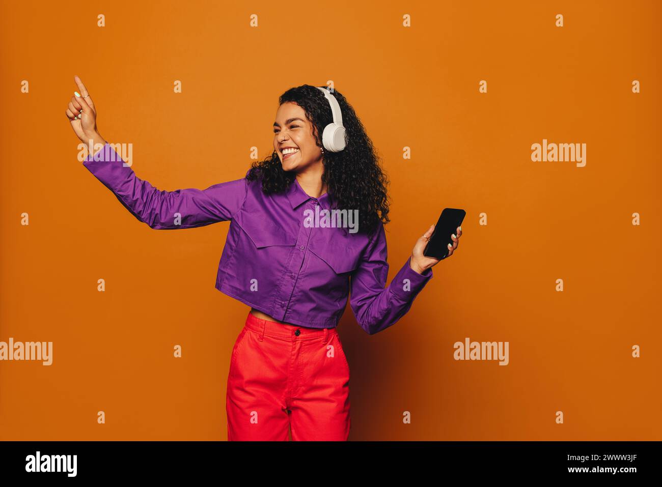 Fröhliche Frau in lässiger Kleidung tanzt zu Musik, Kopfhörer an, hält ein Handy. Leuchtende Farben und ein orangener Hintergrund sorgen für noch mehr Spaß. Stockfoto
