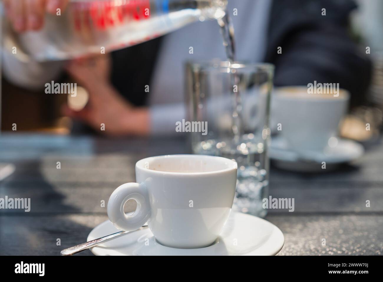 Frau, die Wasser in ein Glas gießt, Tasse Espresso-Kaffee auf schwarzem Tisch im Straßencafé, selektiver Fokus auf Tasse und verschwommenem Hintergrund, Treffen im Café Stockfoto