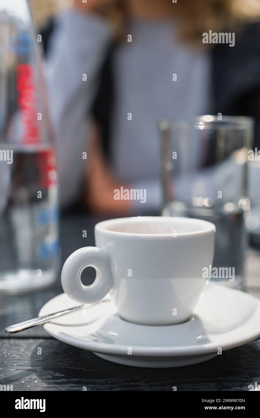 Vertikales Poster mit einer weißen Tasse Espresso auf einem schwarzen Tisch in einem Straßencafé, selektiver Fokus auf die Tasse und unscharfer Hintergrund, Treffen in A Stockfoto
